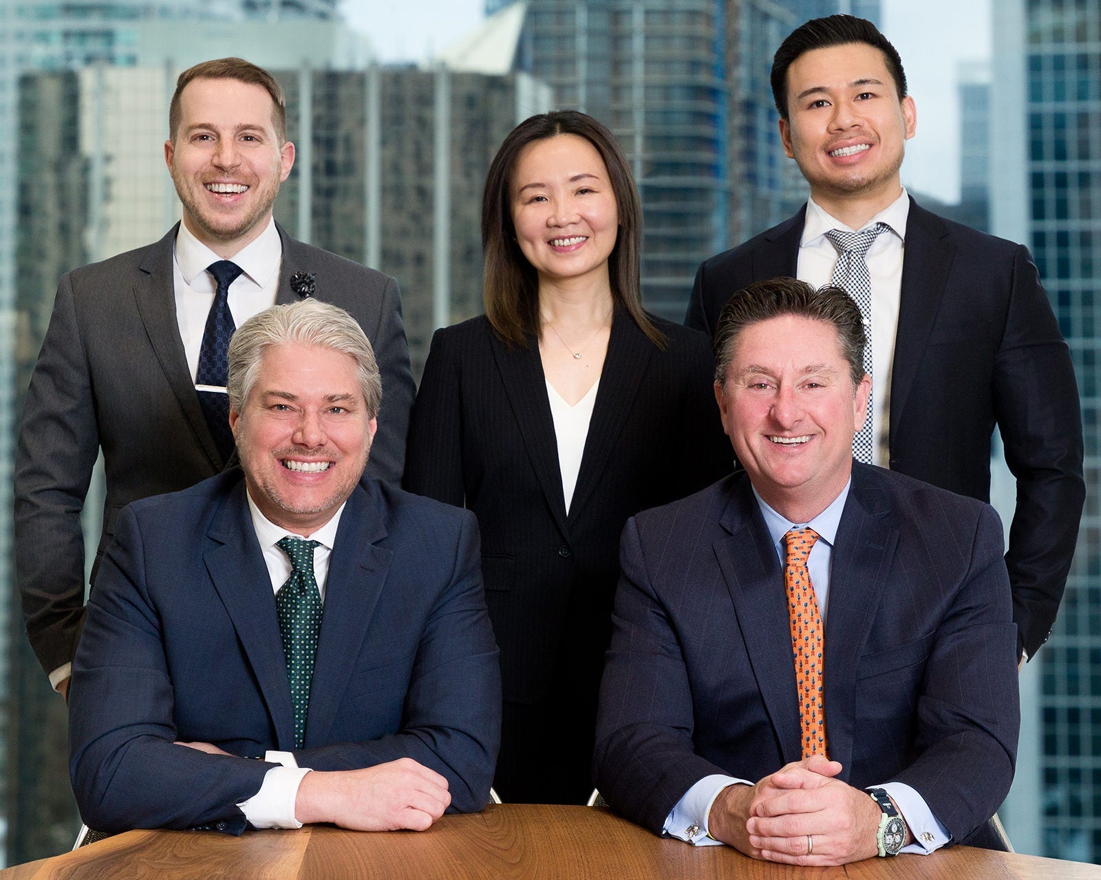 Five colleagues at a boardroom table, two seated and three standing, with a city skyline behind them