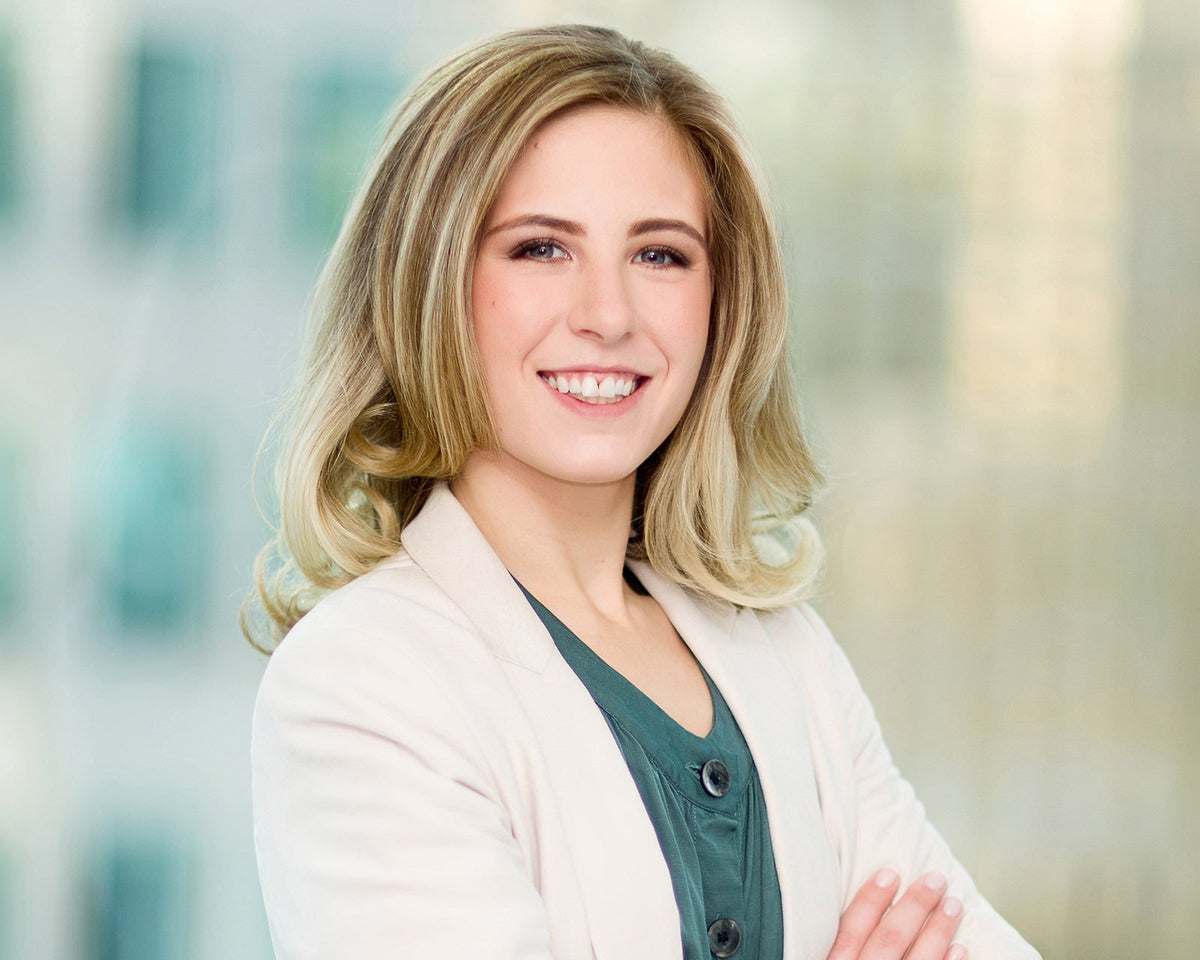 Smiling professional with blonde wavy hair in a white blazer, arms crossed, corporate headshot with a softly blurred office background