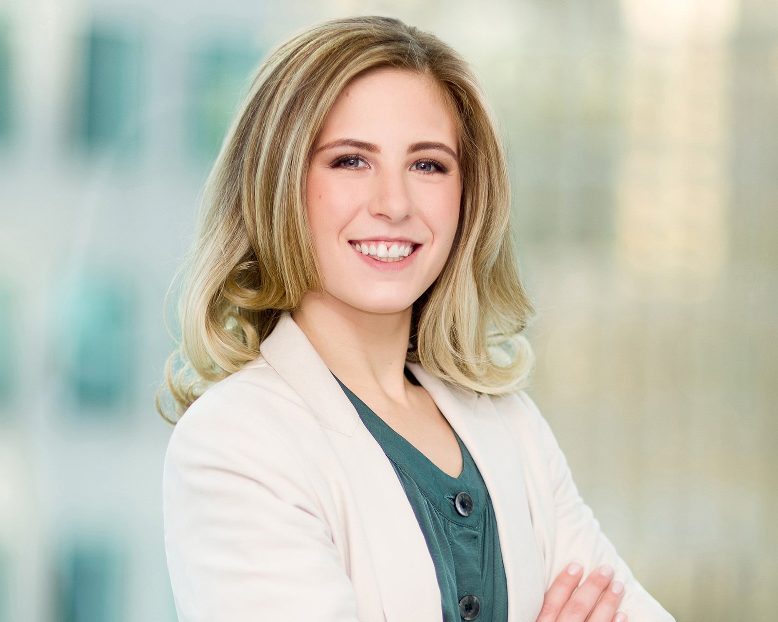 Smiling professional with blonde wavy hair in a white blazer, arms crossed, corporate headshot with a softly blurred office background