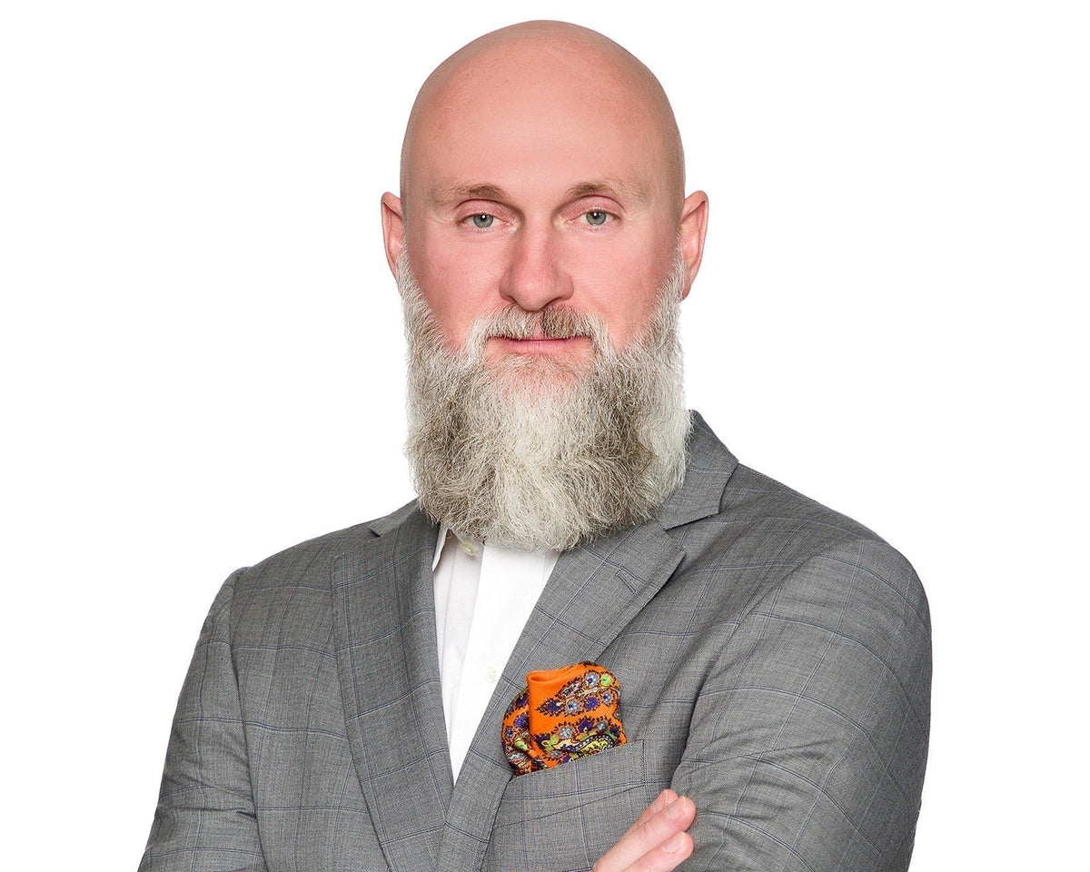 Executive with a full grey beard in a grey check suit and orange patterned pocket square, on a white background.